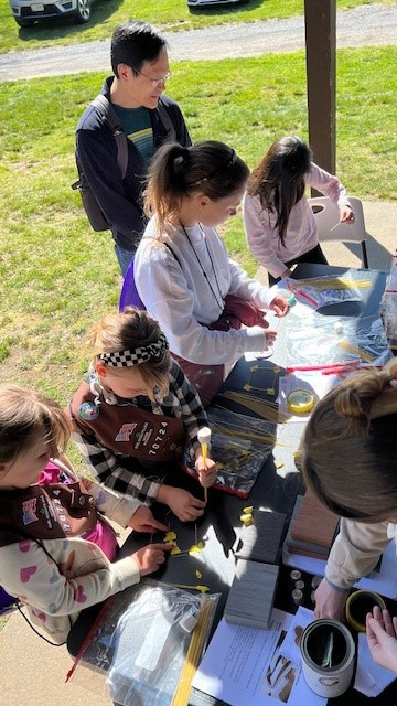 A group of young girls in Brownie uniforms work on a craft project at an outdoor table, enjoying a sunny day in a grassy area. An adult stands nearby smiling, as the sense of community shines through their shared creativity.