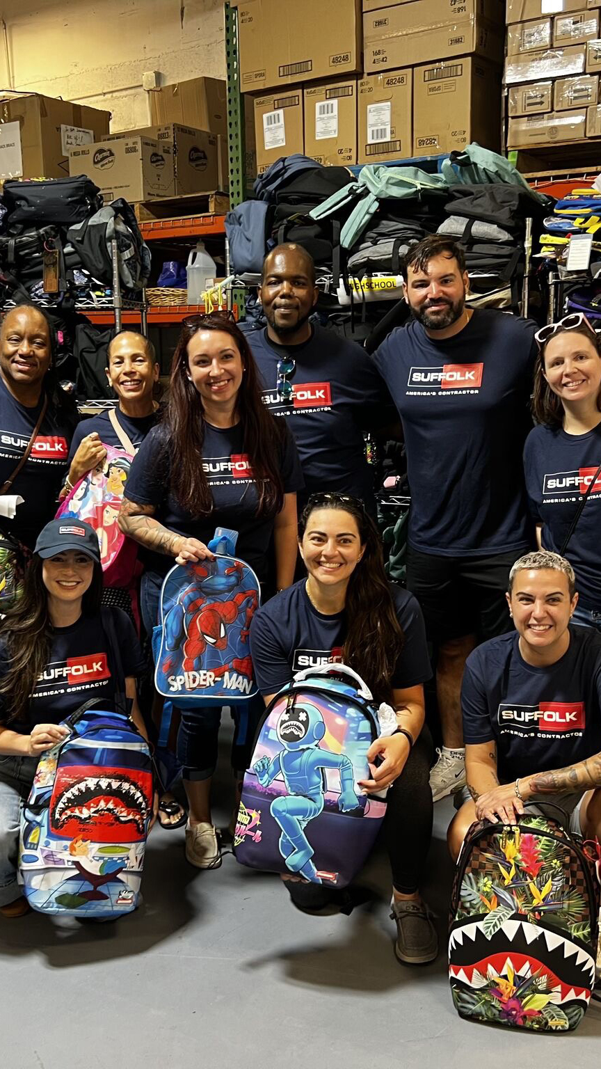 A group of nine smiling people pose in a storage room, proudly holding colorful, cartoon-themed backpacks. Wearing matching SUFFOLK shirts, they showcase their strong sense of community amid shelves full of supplies and boxes.