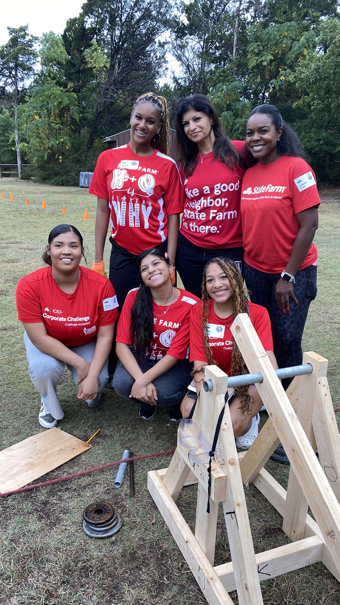 Six women in red State Farm shirts smile outdoors, standing and kneeling around a small wooden catapult on a grassy field with trees in the background. Their cheerful thumbs up reflect their strong sense of community spirit.