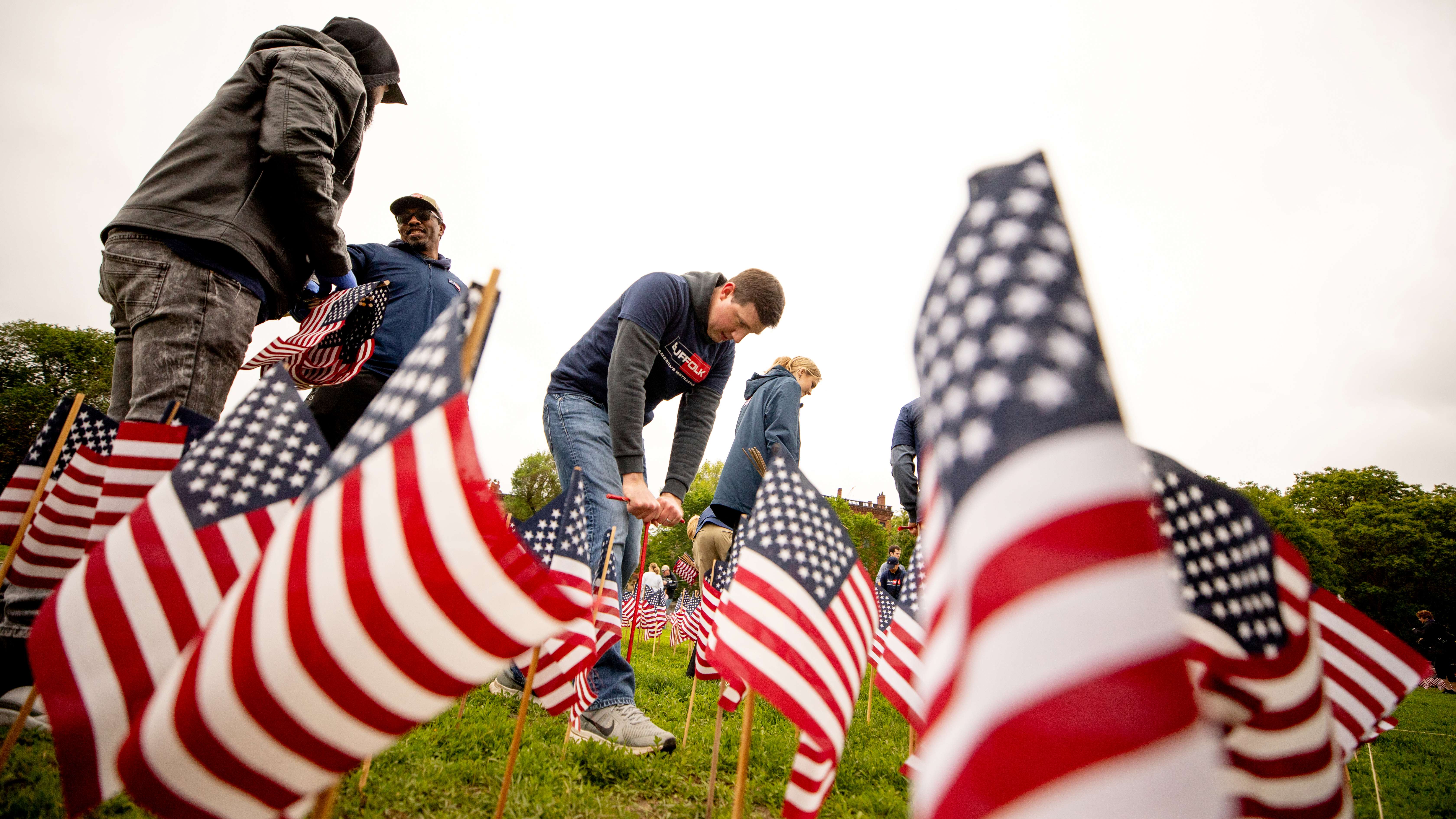 Several people from the community plant small American flags in the grass on a cloudy day, participating in a memorial or patriotic event. The flags are prominent in the foreground, with volunteers working together in the background.