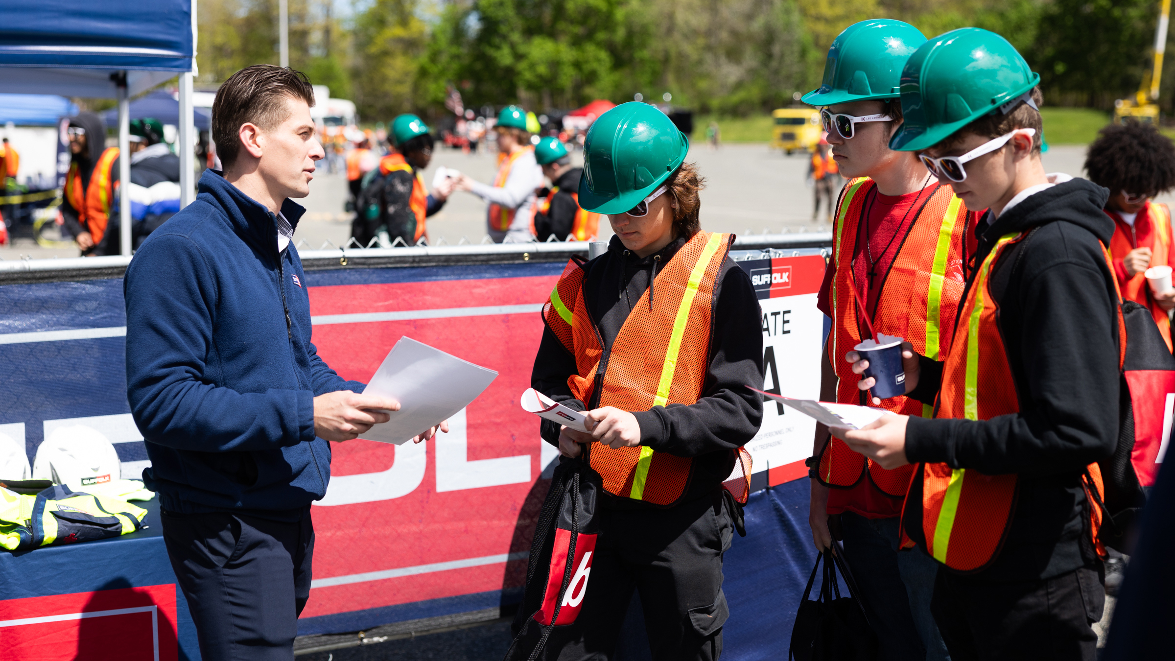 A man in a blue jacket talks to three people wearing green hard hats, orange safety vests, and sunglasses. They are holding papers and standing outside near a construction event area focused on community engagement.