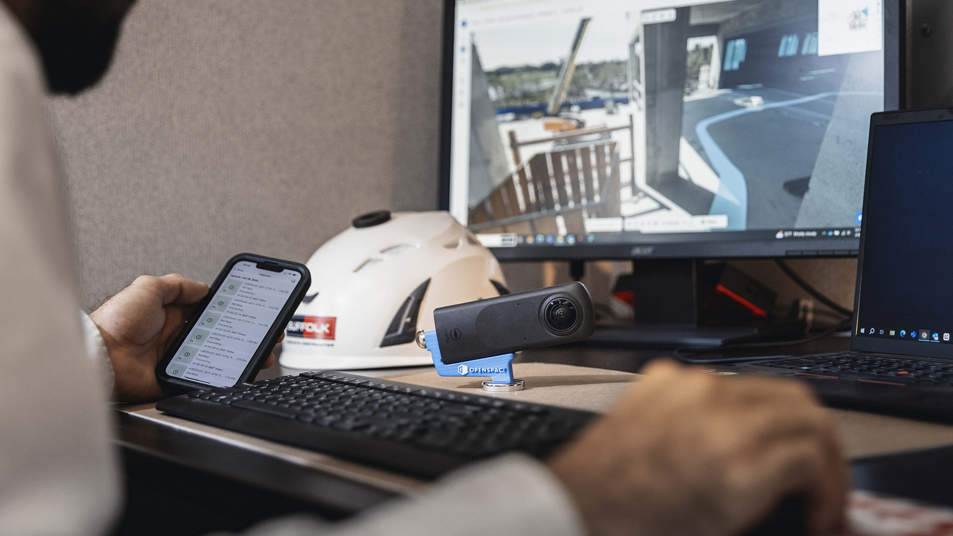 A person sits at a desk using a smartphone and computer, with a helmet, webcam, keyboard, and two monitors showing construction site images in the background.