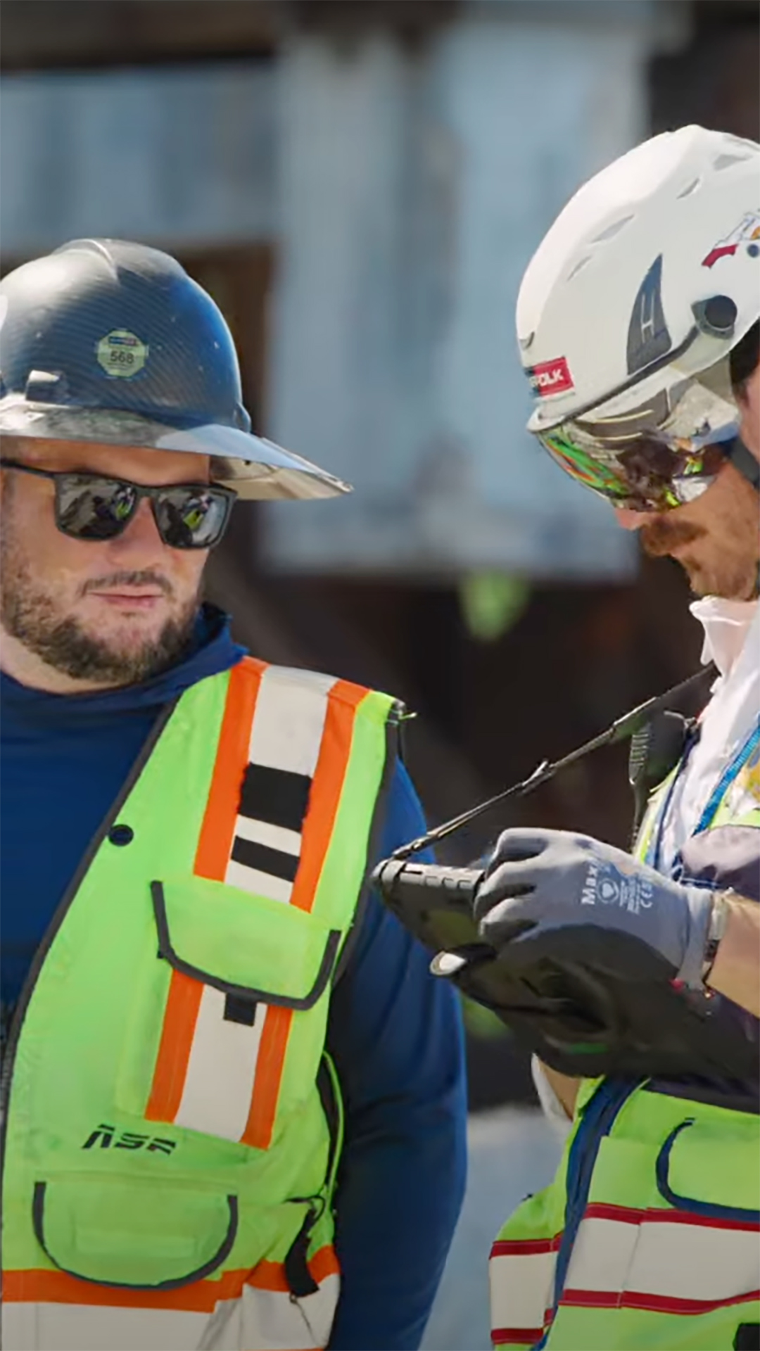 Two construction workers wearing safety vests, helmets, and sunglasses stand outdoors. One is looking at a handheld device that the other is holding, likely discussing or checking something work-related.