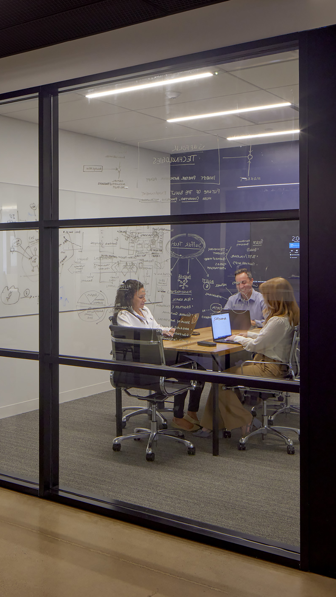 Three people sit around a table in a glass-walled conference room, discussing work with laptops and documents. The walls are covered with diagrams, charts, and notes written in white marker.
