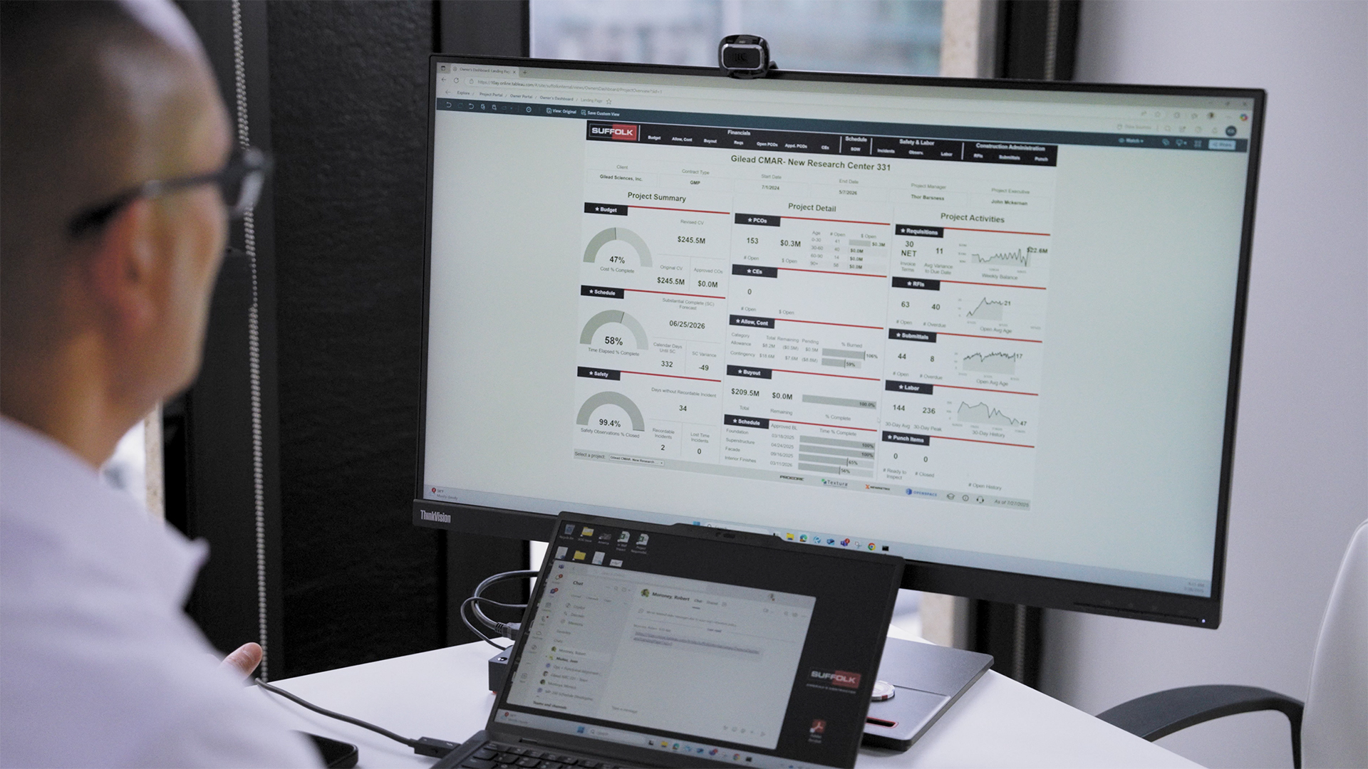 A person sits at a desk viewing project data and charts on a large monitor, with a laptop displaying an email open beside it. The setting is a modern office with large windows in the background.