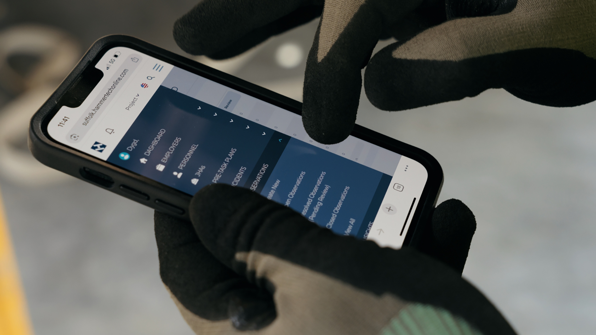 A person wearing black and green work gloves uses a smartphone, browsing a website with a dark blue menu displayed on the screen.