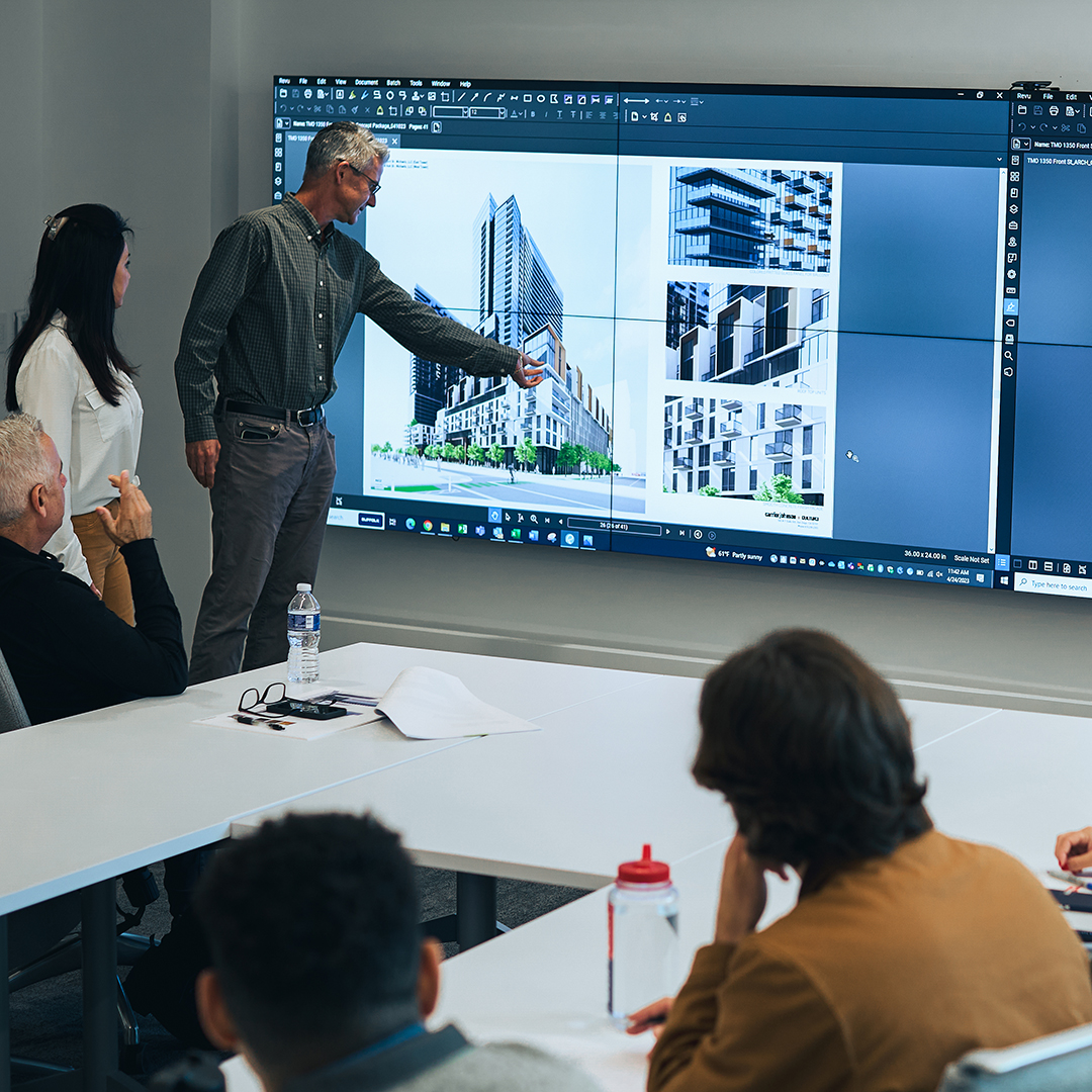 A man points at architectural renderings on a large screen while presenting to a group of people seated at tables in a modern conference room.