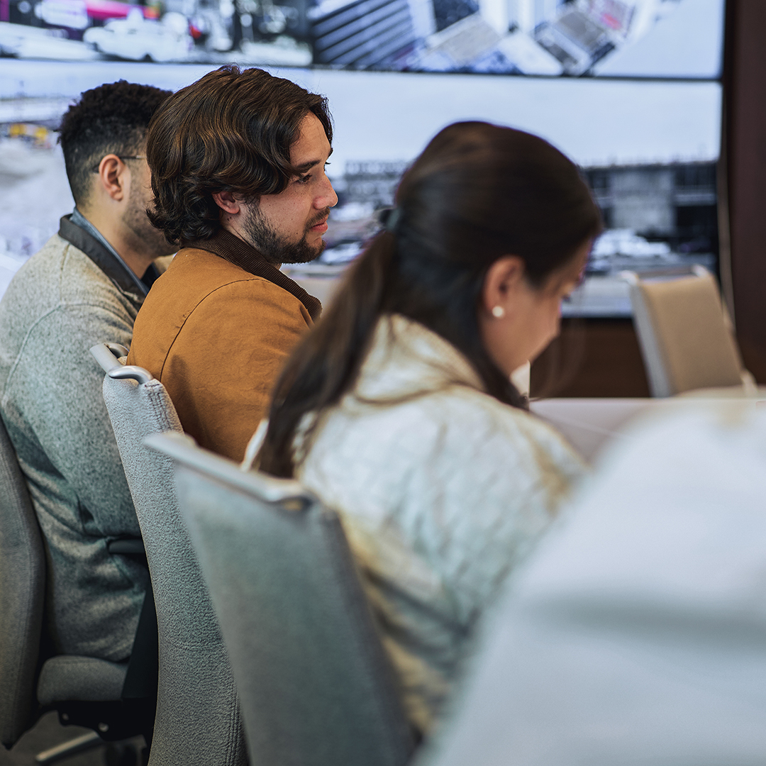 Three people sit in a conference room, facing forward and listening attentively. Large display screens with graphics and images are visible in the background. The focus is on a man with brown hair in a brown jacket.