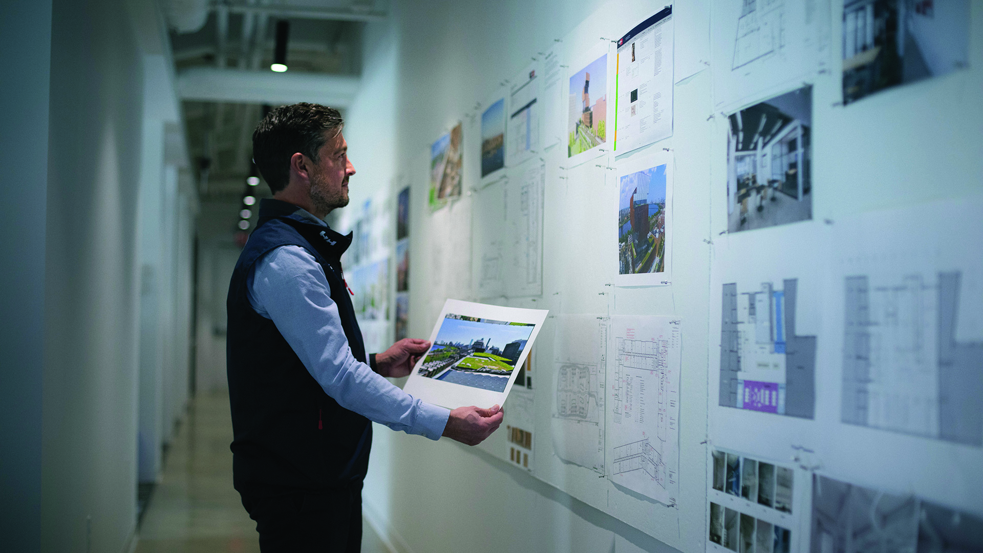 A man stands in a hallway, examining a photo in his hands while looking at a wall covered with architectural plans, blueprints, and photographs.