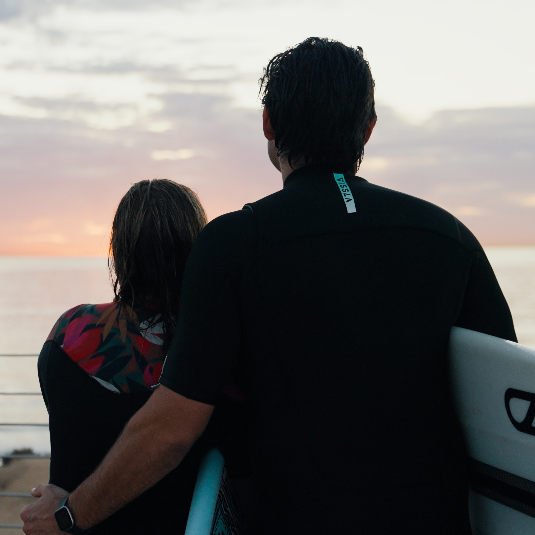 Two surfers in wetsuits stand close together, facing a sunset over the ocean. One person holds a surfboard, enjoying the calming benefits of the peaceful scene and the tranquil connection to nature.