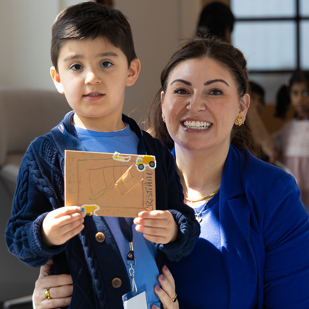 A young boy proudly holds up a handmade craft, showcasing the benefits of creative activities, while standing next to a smiling woman in a blue jacket. They are indoors, with other children blurred in the background.