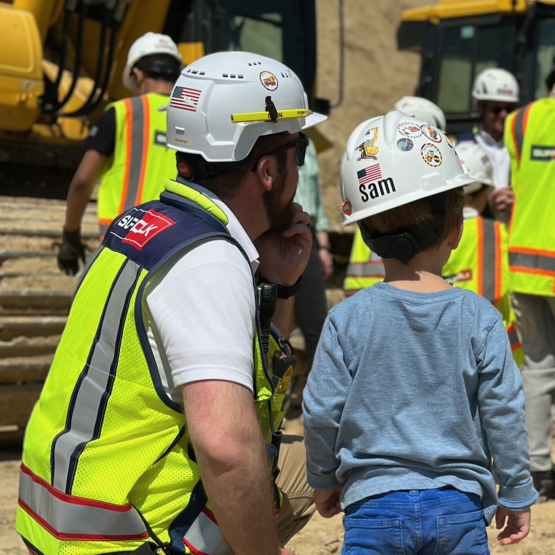 A construction worker kneels beside a young child wearing a hard hat labeled Sam, both observing the benefits of teamwork as workers in safety vests and helmets coordinate near large construction equipment at the work site.