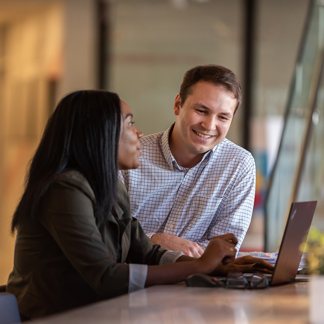 Two people sit at a table, smiling and talking while looking at a laptop, discussing the benefits of their project. The setting appears to be a modern office or coworking space with glass walls in the background.