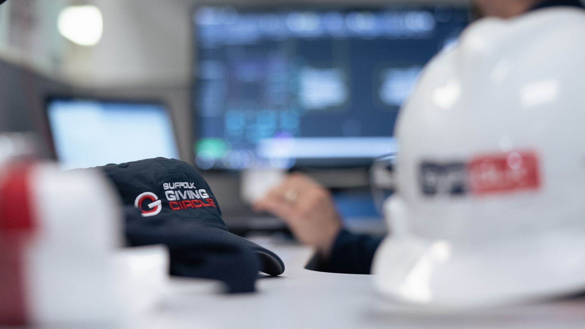 A close-up of a black Suffolk Giving Circle cap and a white construction helmet on a desk, with a blurred person and computer screen in the background.