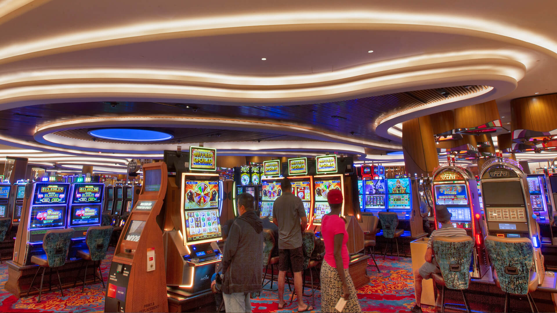 People stand and play at brightly lit slot machines in a colorful casino, one of the most energetic sectors of entertainment, with patterned carpets, curved ceiling lights, and rows of gaming machines. The atmosphere appears lively and vibrant.