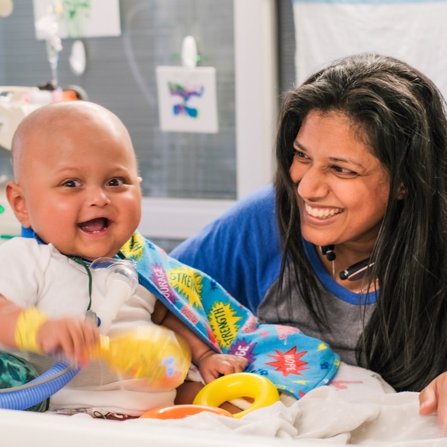 A smiling baby with medical equipment and colorful toys is lying in a hospital bed, next to a woman who is also smiling warmly at the baby.