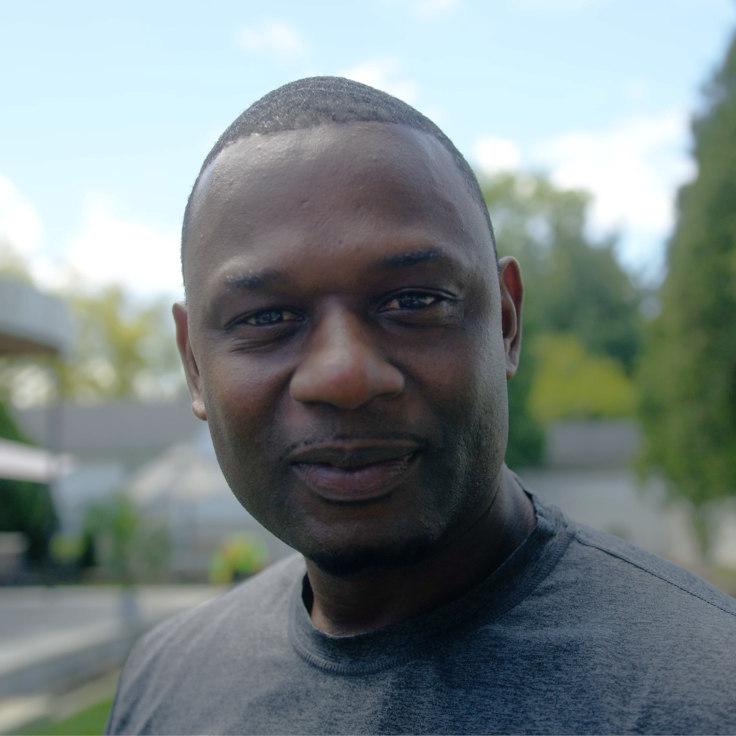 A man with short hair and a close-cropped beard smiles slightly outdoors on a sunny day. He is wearing a charcoal gray T-shirt, with green trees and blurred buildings in the background.