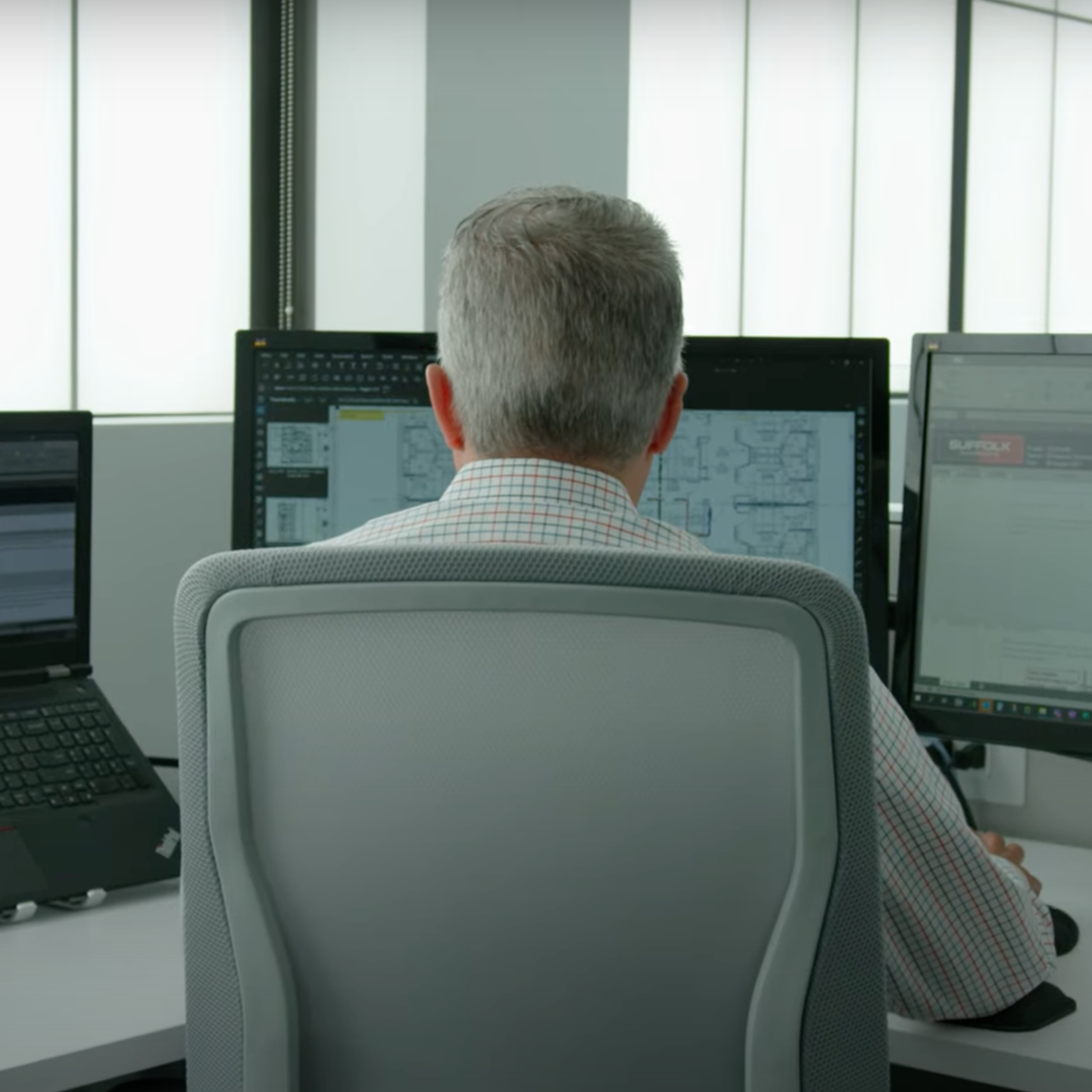 A person with short gray hair sits in an office chair, facing three computer screens displaying documents and diagrams in a modern workspace.