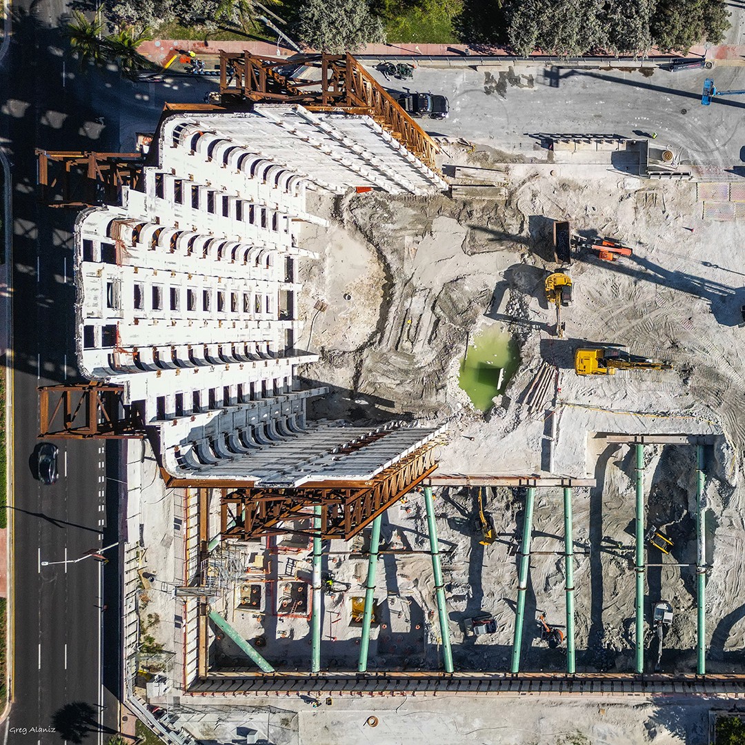 Aerial view of a building under partial demolition, with only the front facade remaining upright. Construction equipment and debris are visible around the site, with exposed foundations and a small pool of green water.