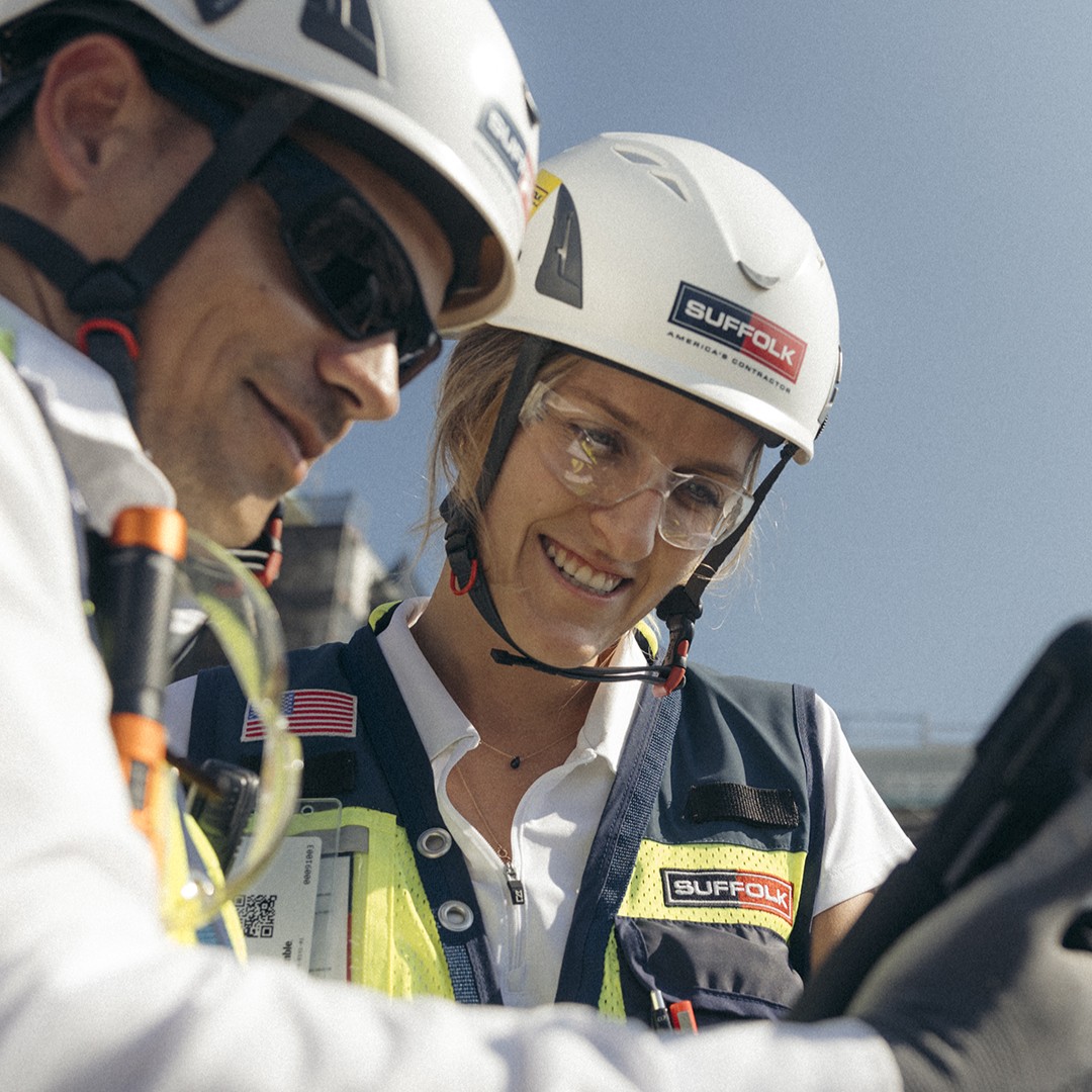 Two construction workers wearing safety helmets, glasses, and vests are looking at a tablet and smiling under a clear blue sky. The worker in focus is a woman with a Suffolk logo on her helmet and vest.