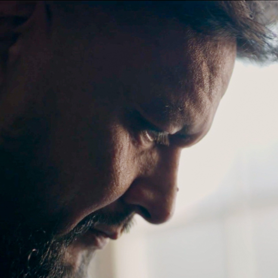 Close-up of a man’s face in profile, looking down with a serious or thoughtful expression. Soft light illuminates part of his face, highlighting the texture of his skin and beard.