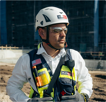 A construction worker wearing a white helmet, safety glasses, gloves, and a reflective vest with badges and tools stands outdoors at a construction site, helping to build a new home while looking off to the side.