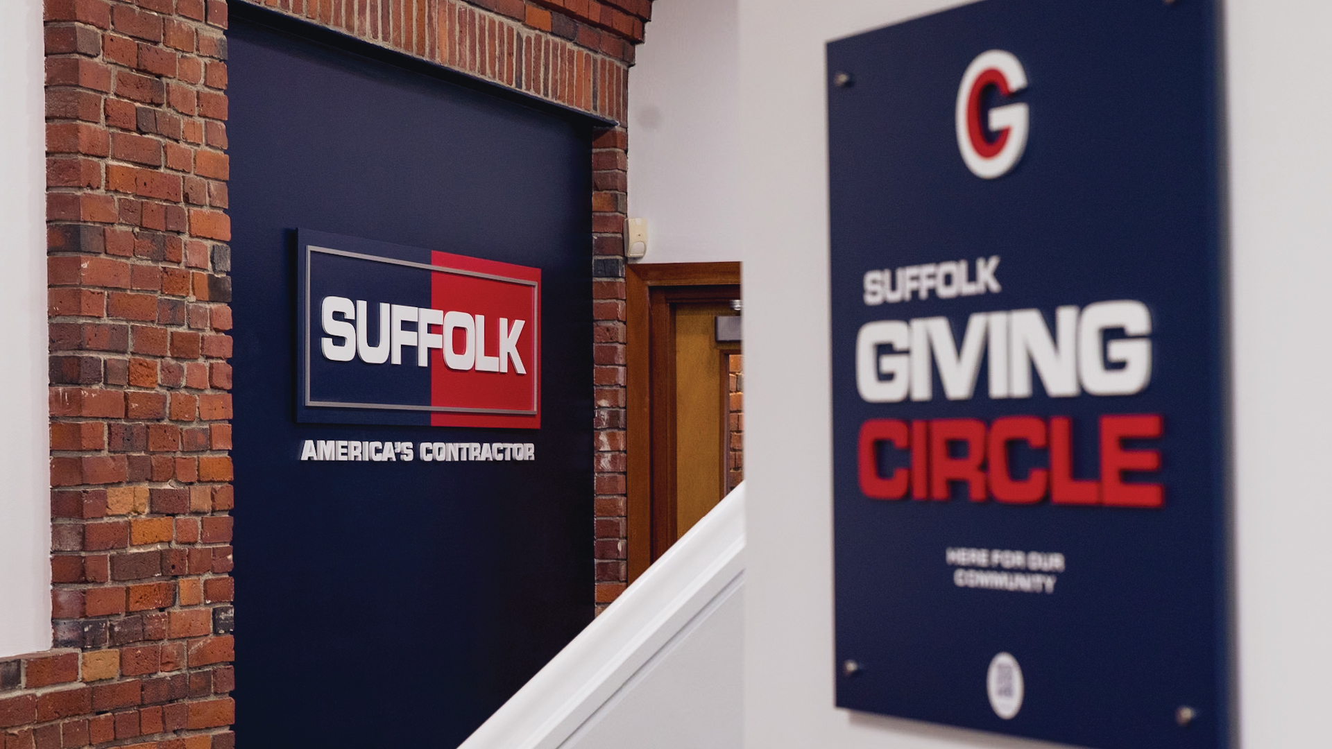 A hallway with exposed brick walls and two signs. One sign reads SUFFOLK AMERICAS CONTRACTOR and another nearby reads SUFFOLK GIVING CIRCLE HERE FOR OUR COMMUNITY with a red and blue color scheme.