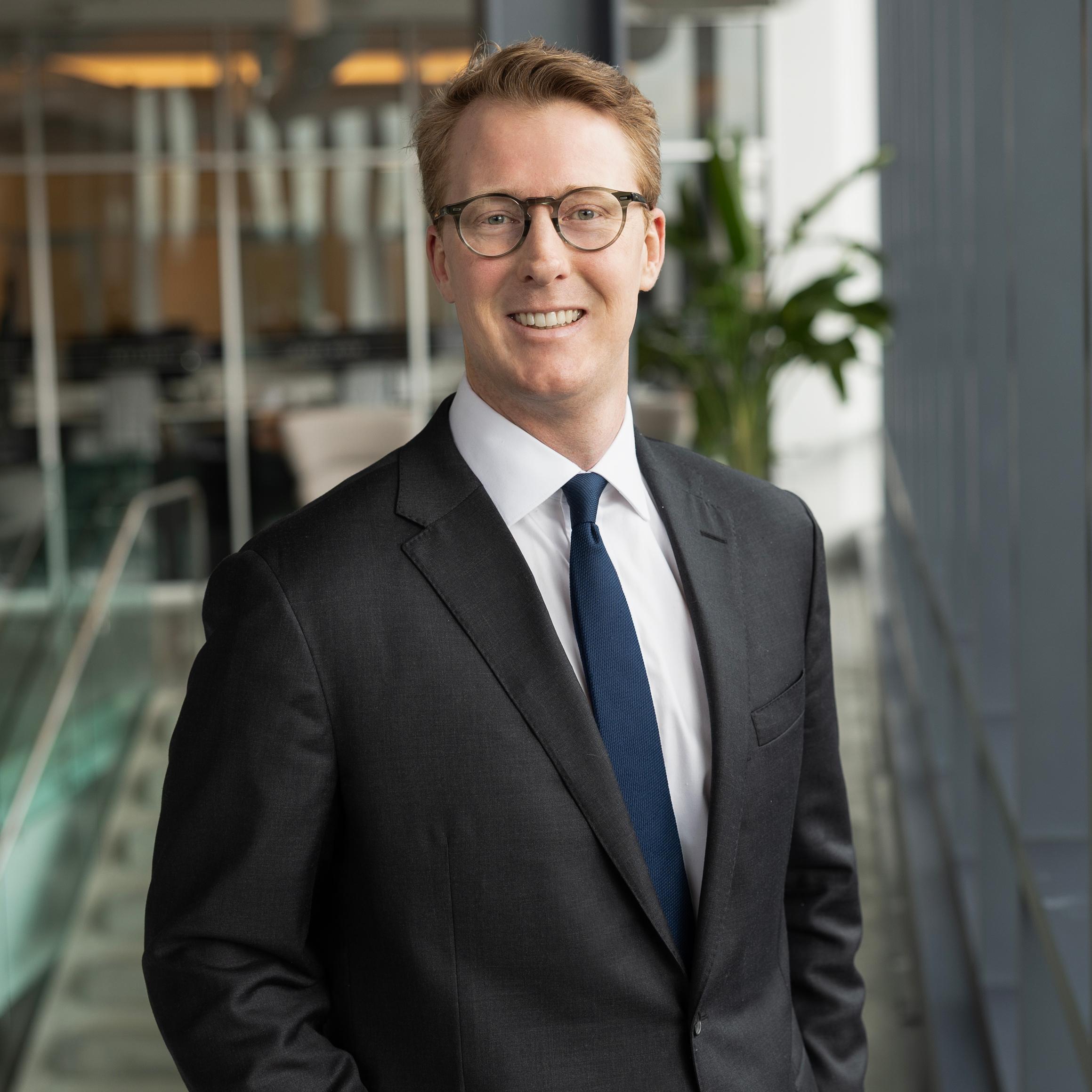 A man wearing glasses, a dark suit, white shirt, and navy tie smiles while standing indoors in a modern office space with glass walls and a potted plant.