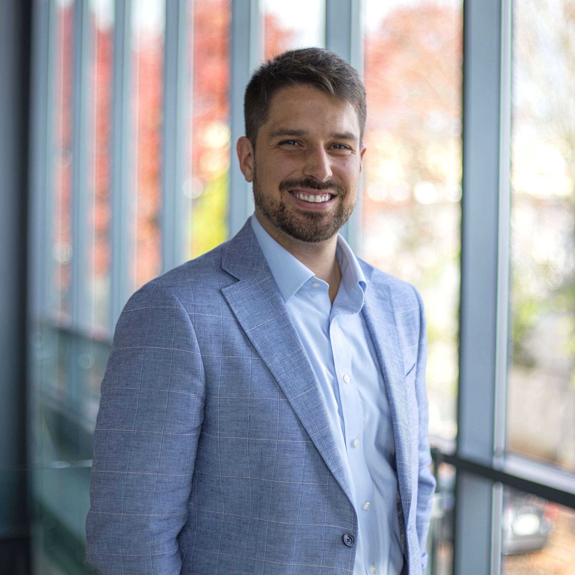 A man with short brown hair and a beard, wearing a light blue suit jacket and white shirt, stands smiling in front of large windows with blurred outdoor scenery in the background.