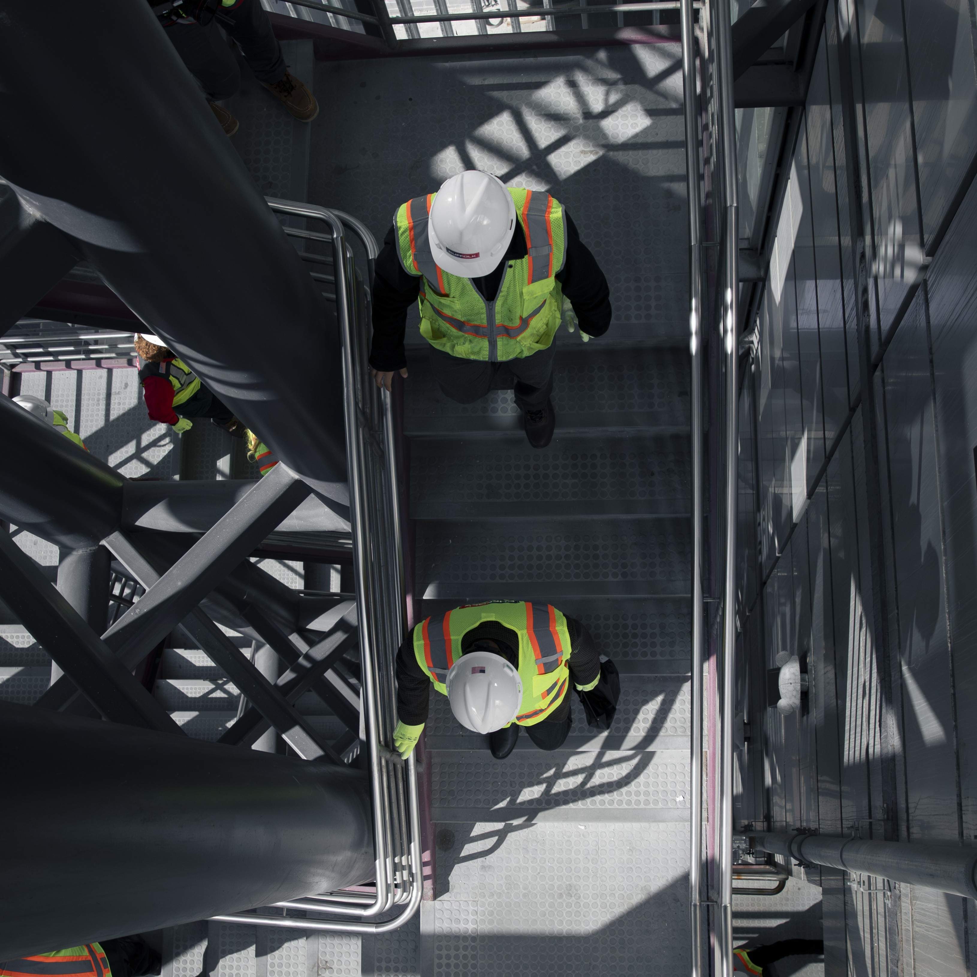 Two construction workers in safety vests and hard hats walk up a metal staircase inside an industrial building, viewed from above. Shadows from the structure create a geometric pattern, highlighting the sense of community in their teamwork.