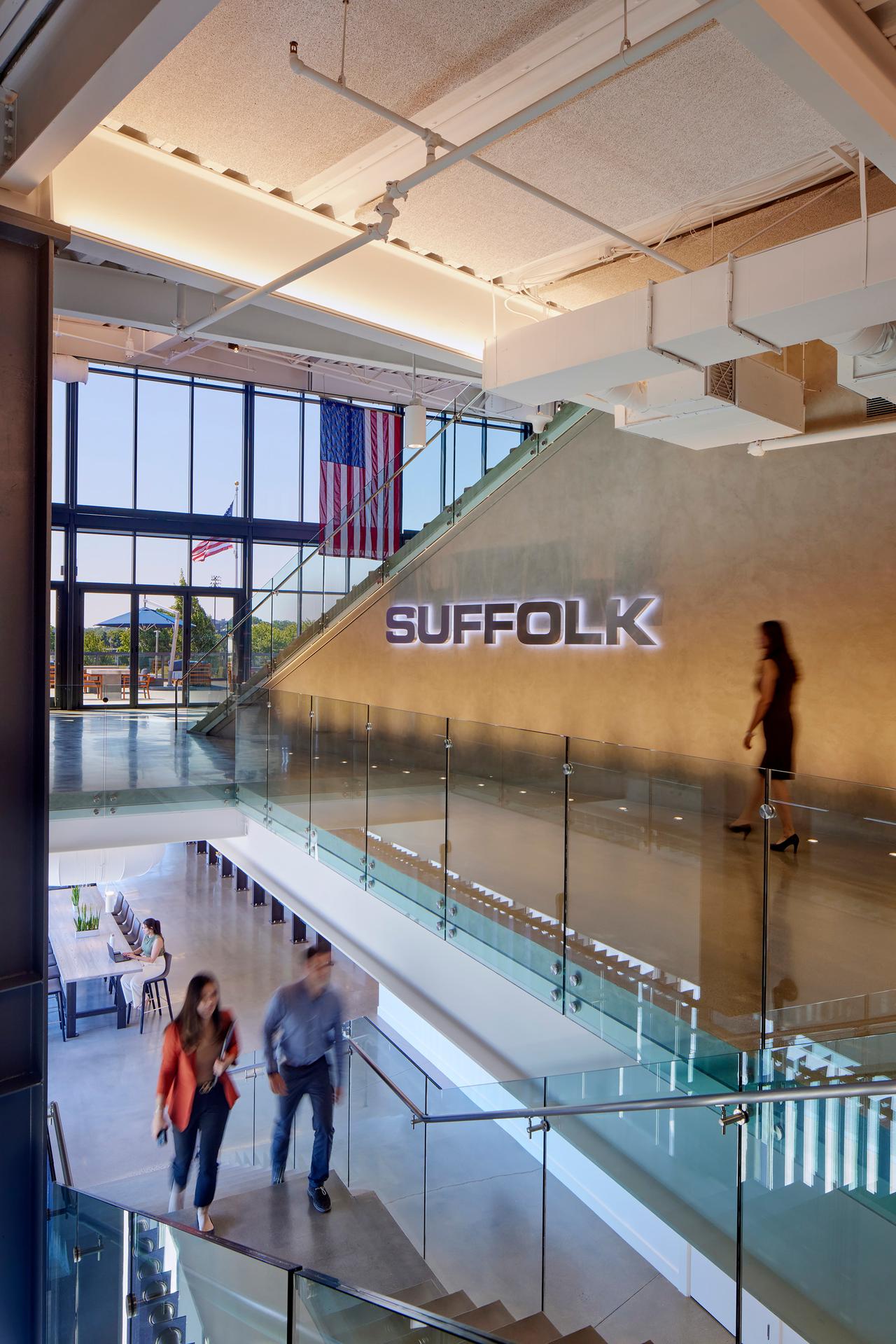 A modern office interior showcases The Suffolk System with glass railings, a large SUFFOLK sign on the wall, people walking on stairs, and American flags visible through tall windows in the background.