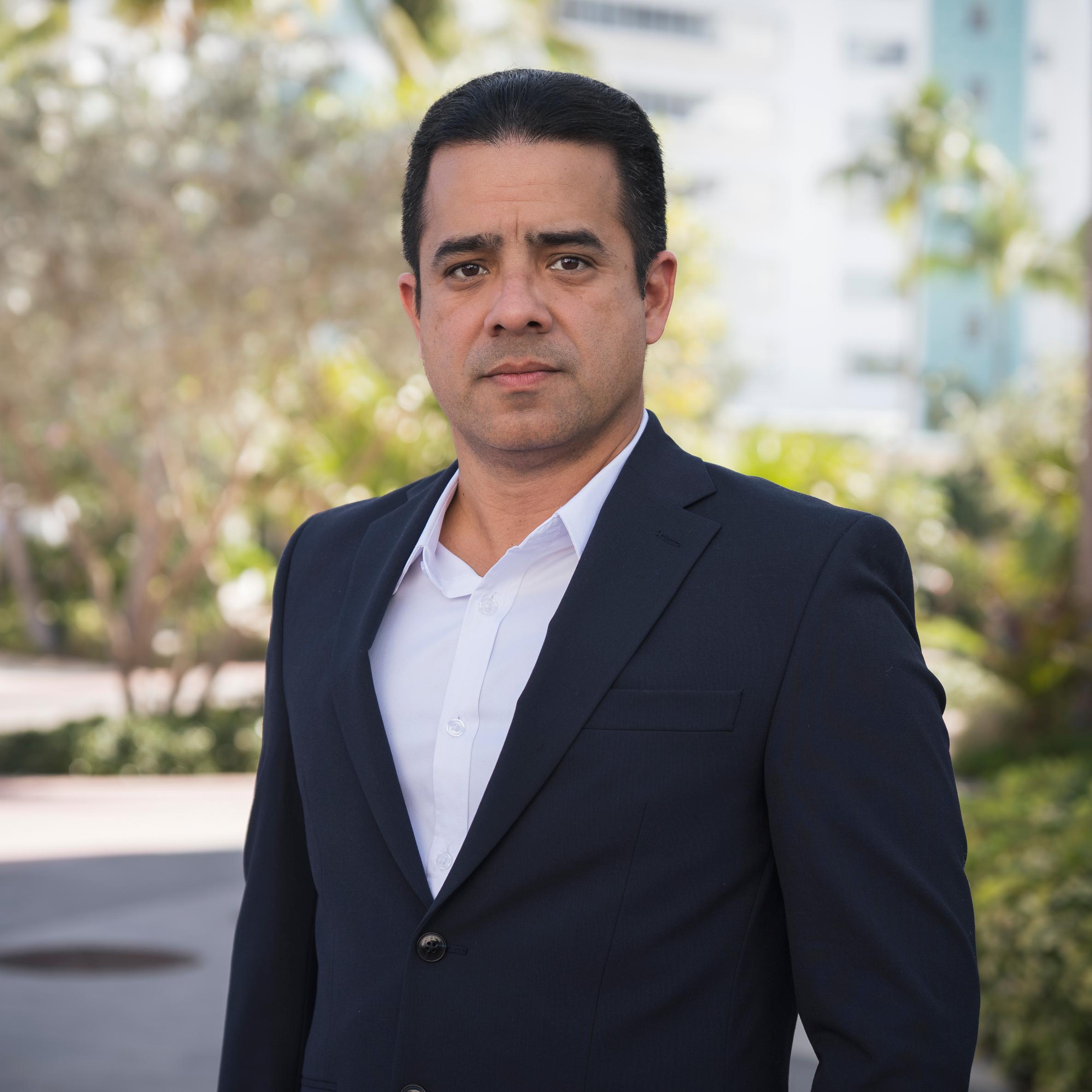 A man with short dark hair wearing a navy suit and white shirt stands outdoors. Trees and a building are visible in the blurred background. He looks directly at the camera with a neutral expression.