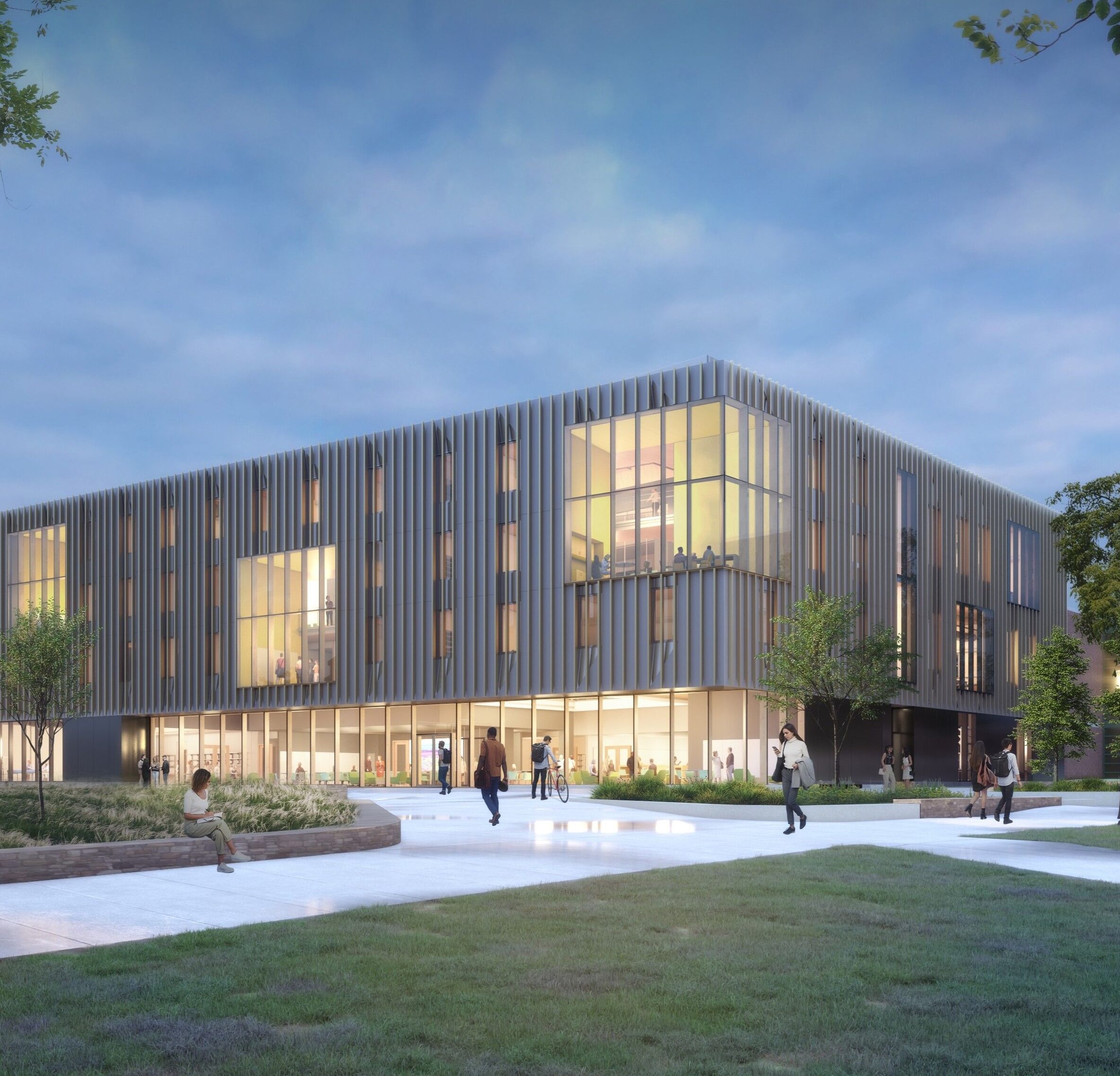 A modern, three-story building with large windows is lit up at dusk, featuring vertical metal siding. People walk along paths and grassy areas in front of the building, surrounded by trees and landscaping.
