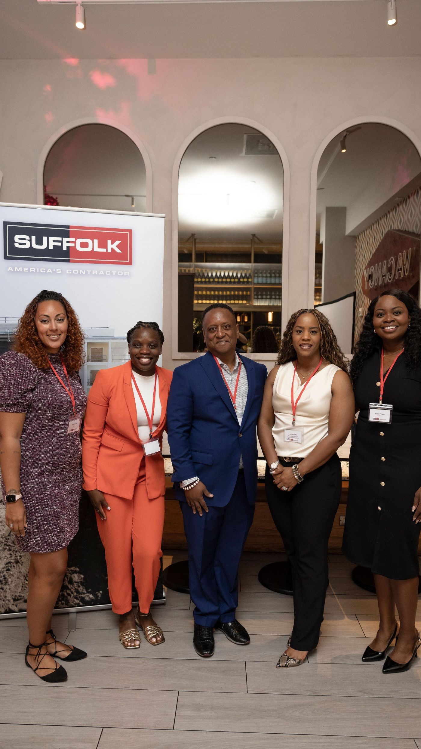Five professionally dressed people stand indoors in front of a Suffolk construction sign, smiling at the camera. Three women wear dresses, one wears an orange suit, and one man wears a blue suit.