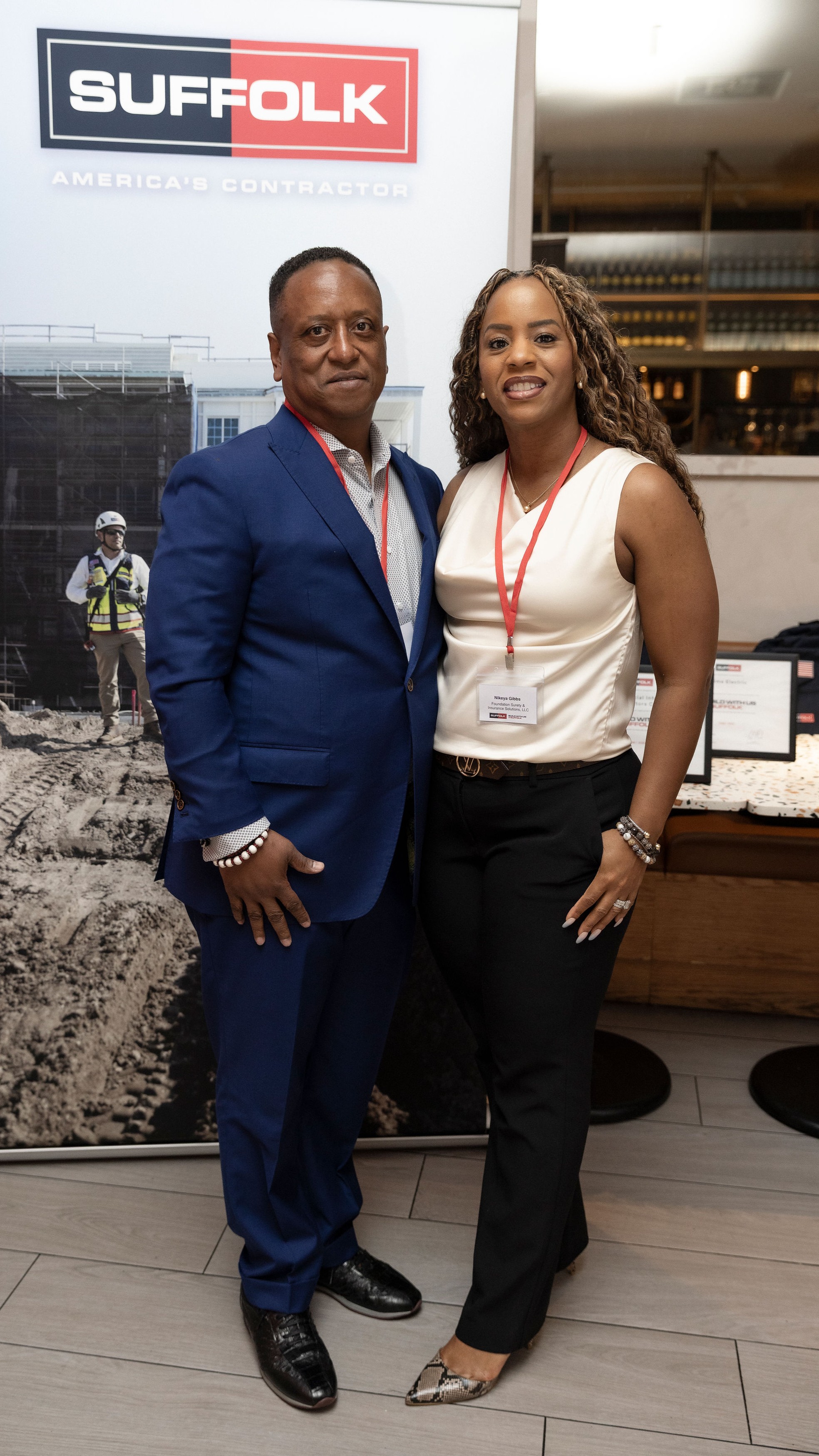 A man in a blue suit and a woman in a white blouse and black pants stand smiling together in front of a Suffolk construction company banner at an indoor event.
