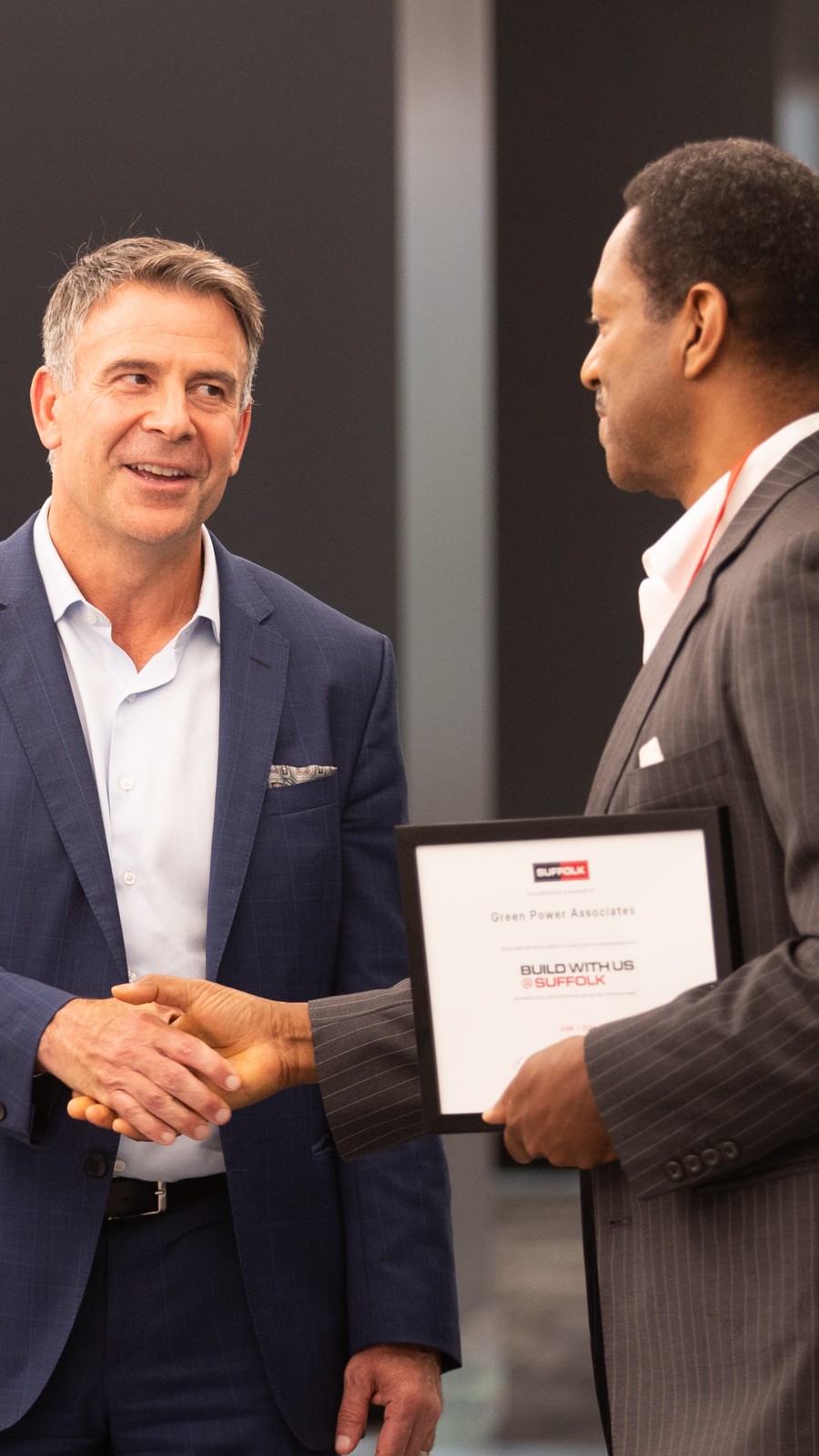 Two men in business attire shake hands. One man smiles while the other holds a framed certificate that reads BUILD WITH US. They appear to be at a professional event or ceremony.
