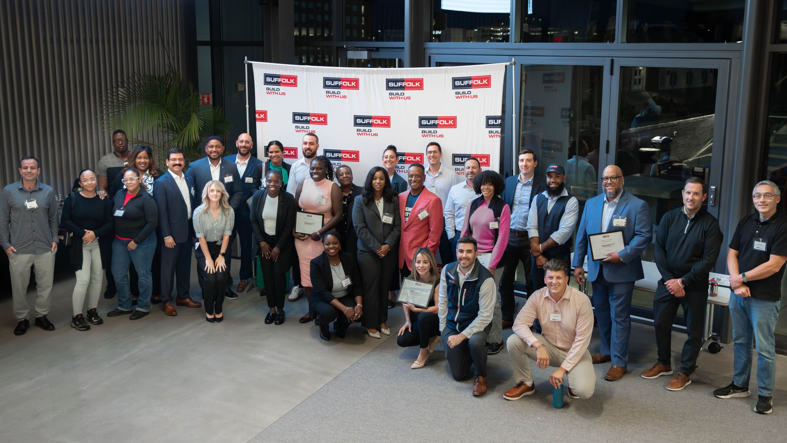 A diverse group of people pose together indoors, some holding certificates, in front of a red-and-white Suffolk Build With Us banner. The group is smiling, and the setting appears professional and celebratory.