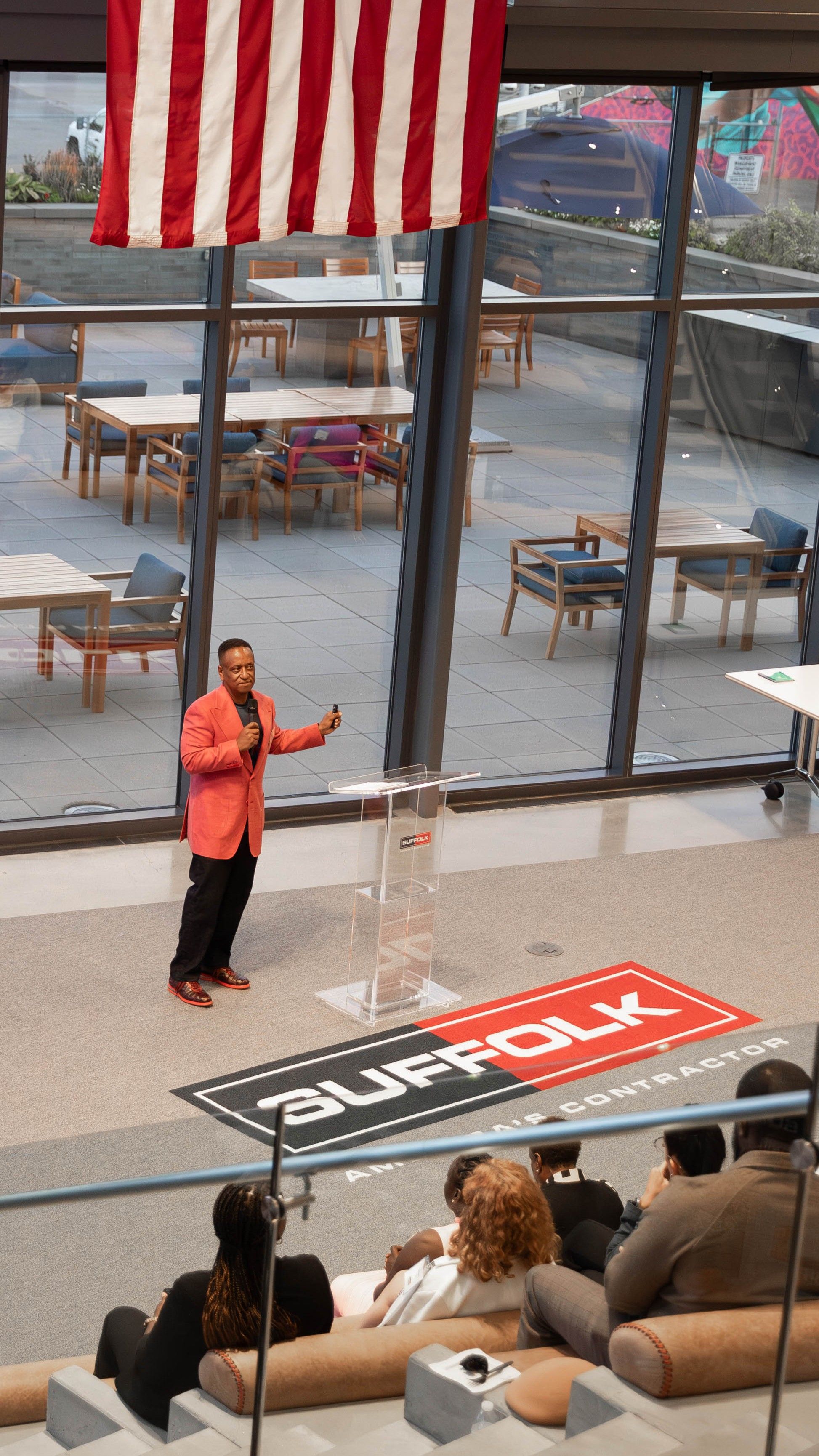 A man in a red jacket speaks at a clear podium in front of an audience inside a modern building, with a large American flag above and the SUFFOLK logo on the floor below him.