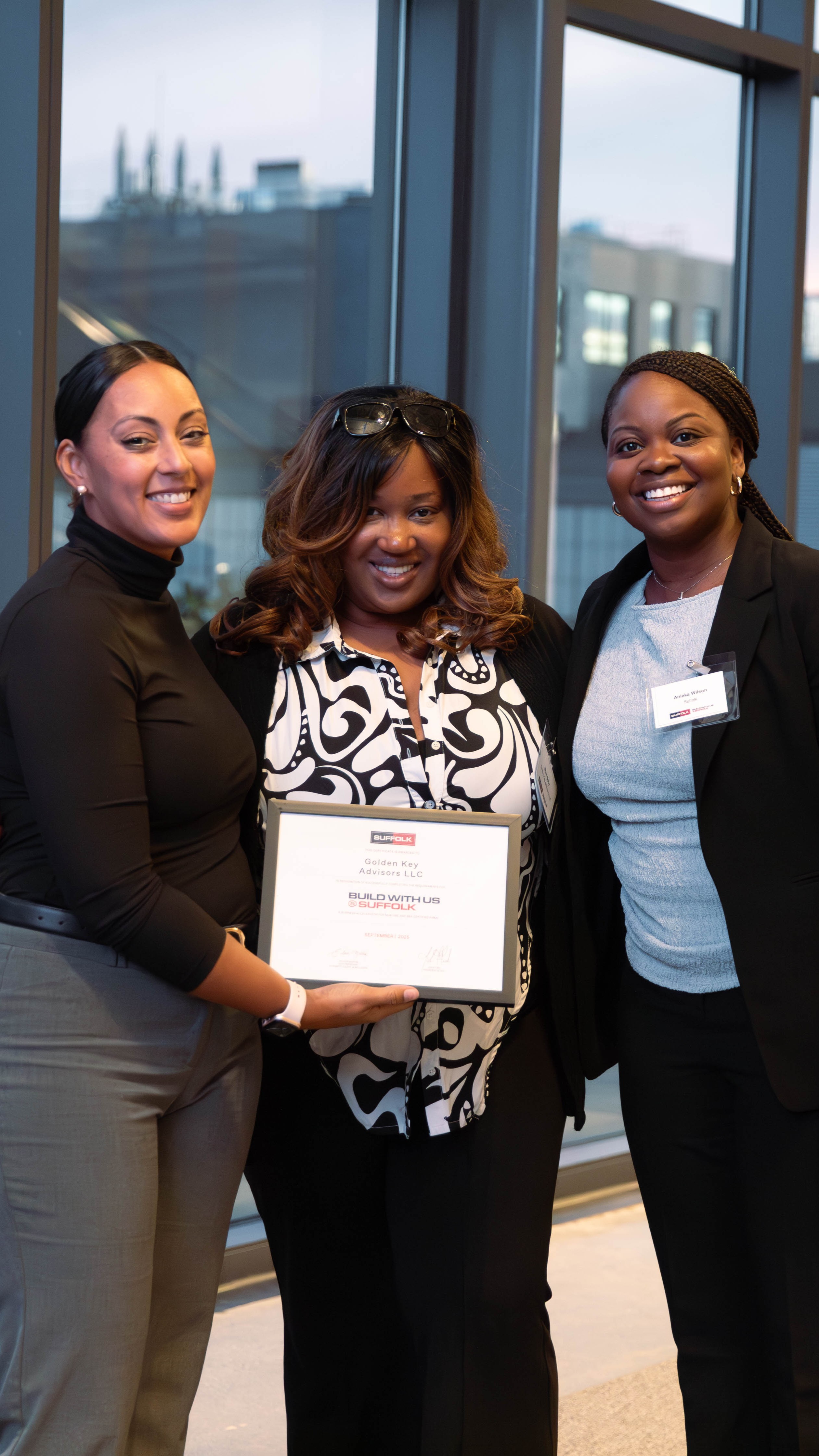 Three women smiling and posing indoors, with the woman in the center holding a certificate. They are dressed in business attire and standing in front of large windows.