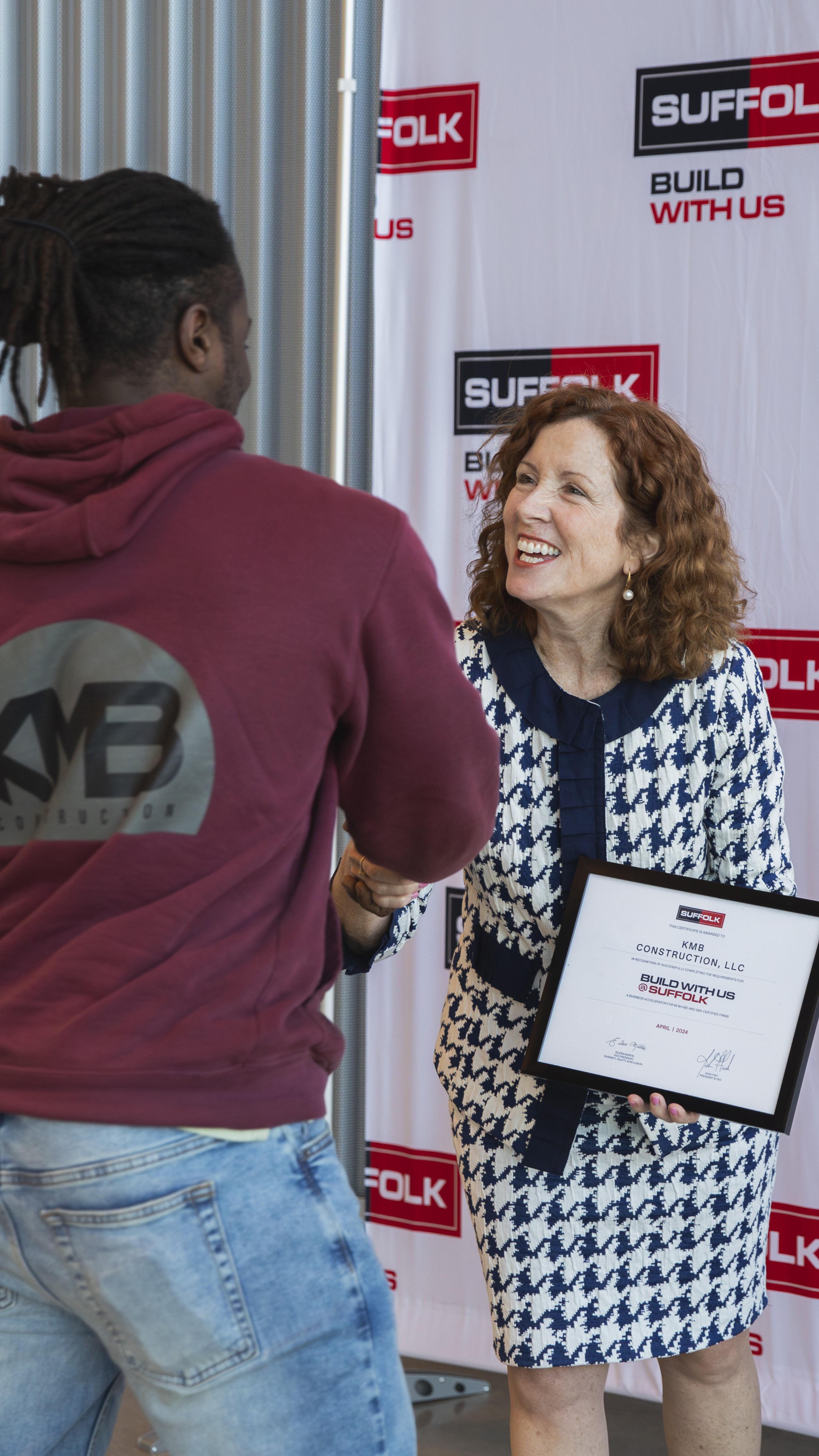 A woman in a blue and white suit smiles and shakes hands with a man in a maroon hoodie while holding a framed certificate, standing in front of a backdrop with SUFFOLK and BUILD WITH US logos.