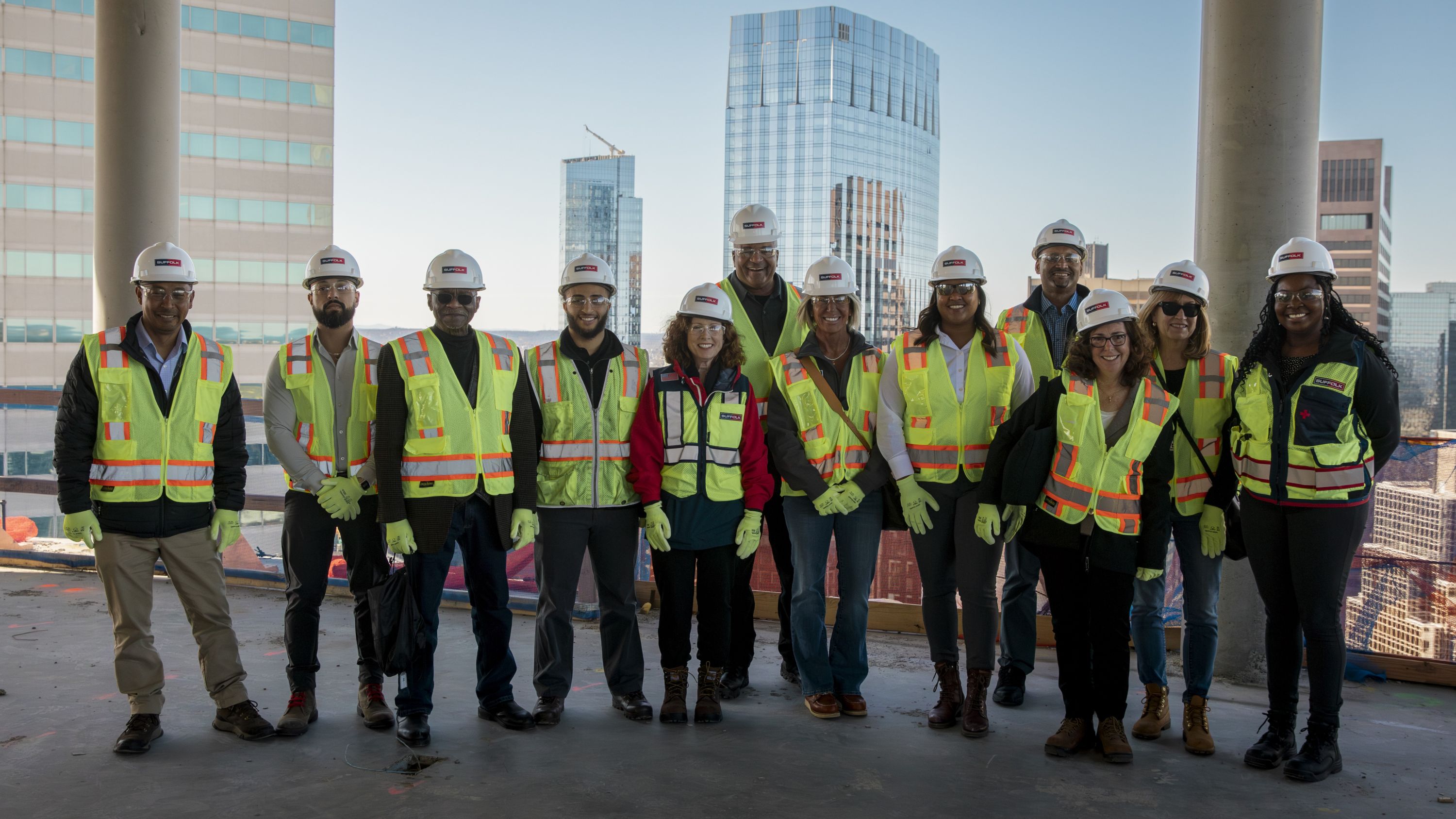 A group of twelve construction workers wearing safety vests, hard hats, and gloves stand side by side on a building site with city skyscrapers in the background.
