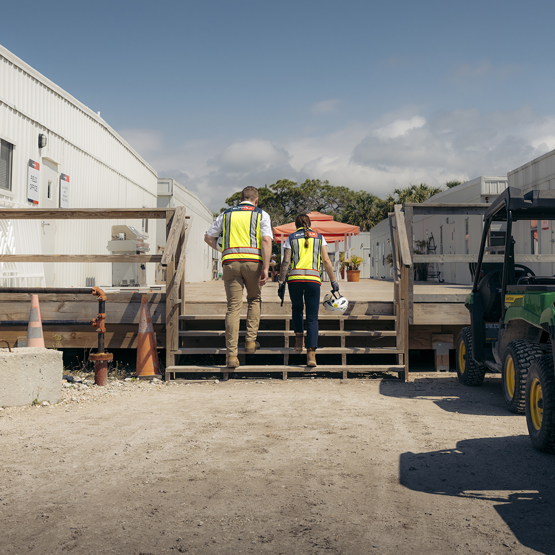 Two construction workers wearing yellow safety vests walk up wooden stairs toward portable office buildings at a job site under a blue sky, highlighting the diverse careers available in the construction industry, with cones and vehicles nearby.