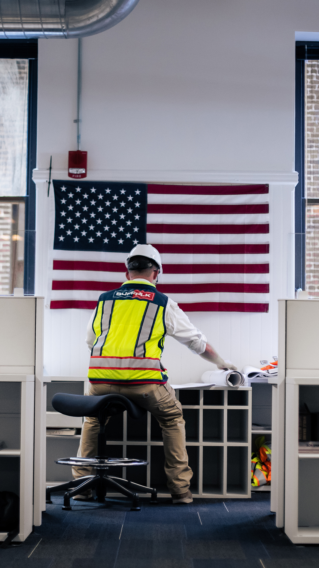 A construction worker pursuing careers in the industry, wearing a safety vest and helmet, reviews blueprints at a desk in an office. An American flag hangs on the wall behind them.