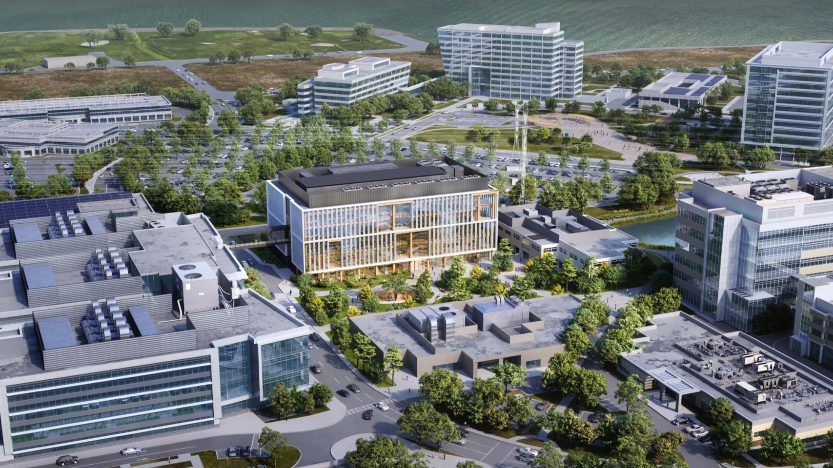 Aerial view of a modern office complex with multiple glass buildings, green landscaping, parking lots, and water in the background. Trees and walkways are visible throughout the area.