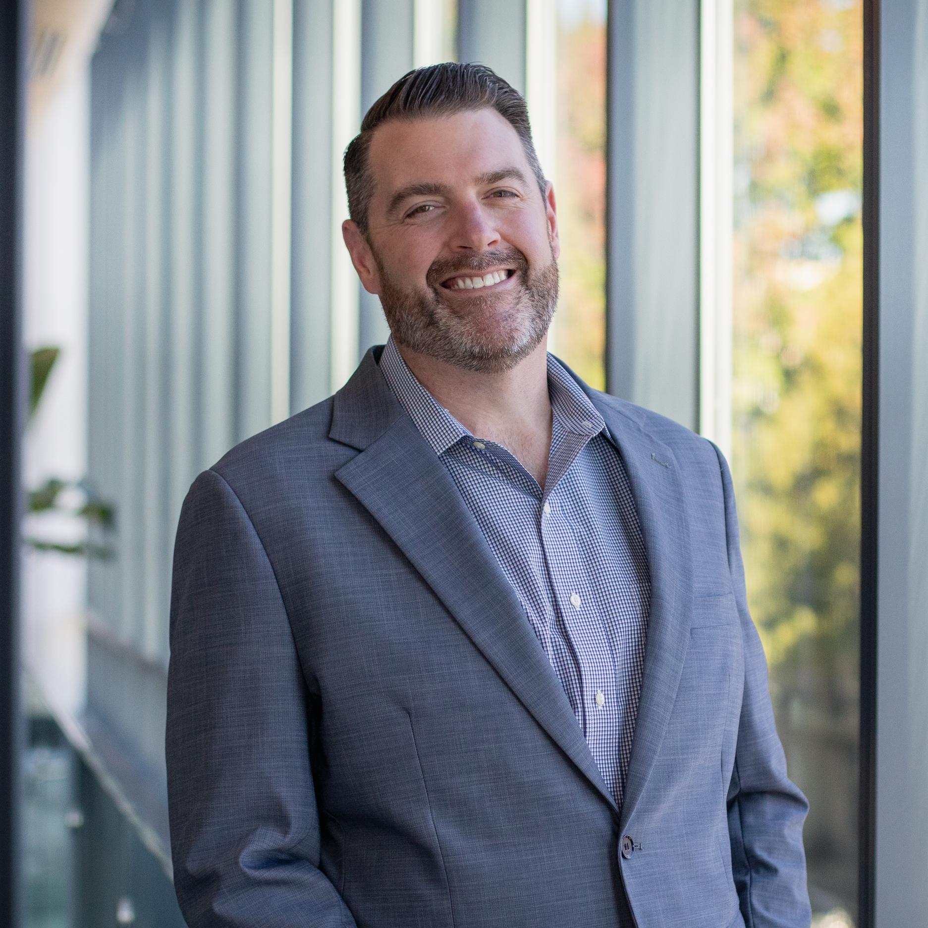 A man with short brown hair and a trimmed beard, wearing a gray suit jacket over a patterned shirt, stands smiling by large windows with sunlight streaming in.