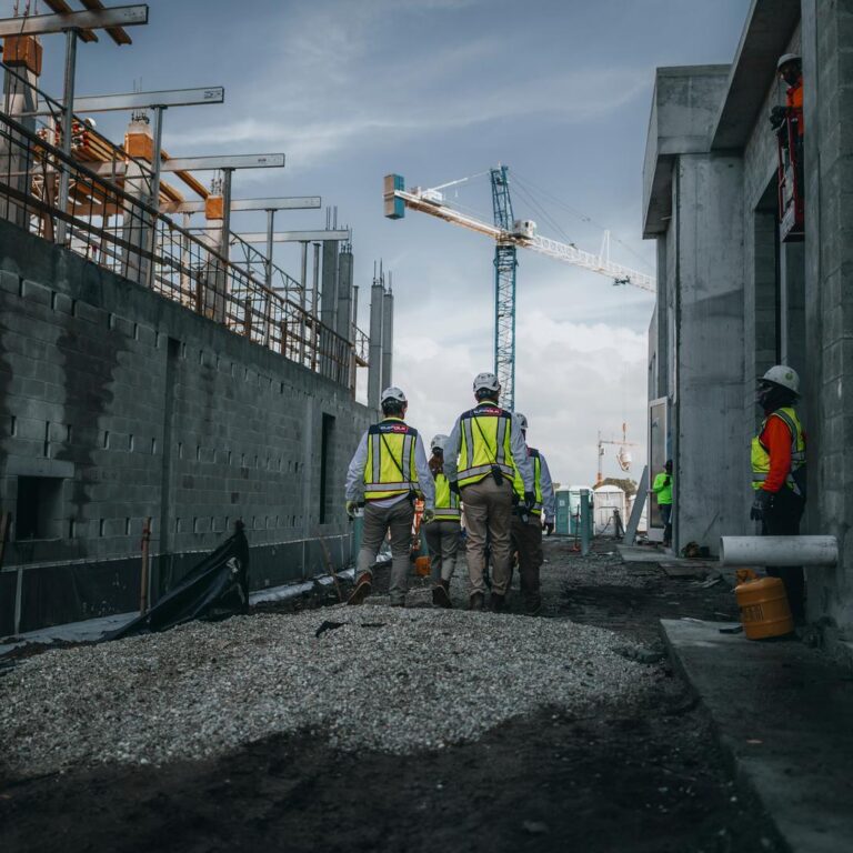 Construction workers wearing safety gear walk through a building site between unfinished walls, with cranes and building materials visible in the background under a cloudy sky.