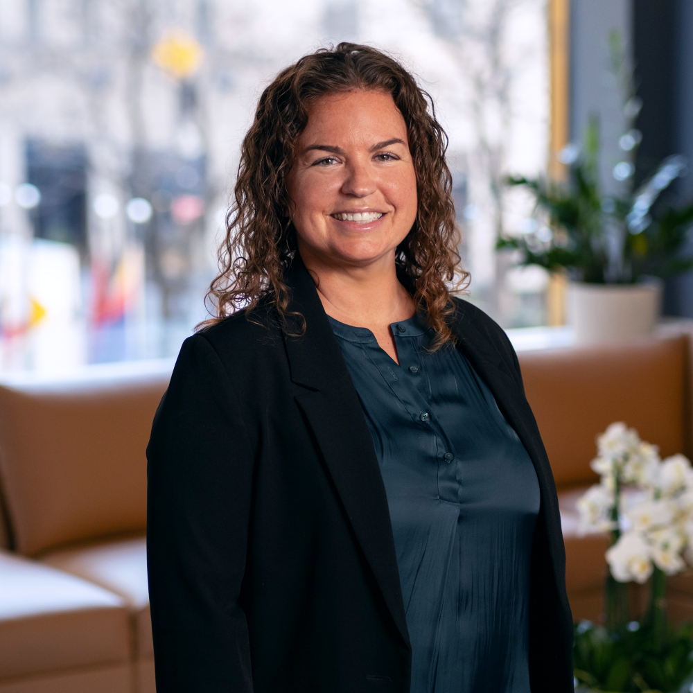 A woman with curly brown hair, wearing a dark blazer and teal blouse, smiles while standing in a modern office with plants and large windows in the background.