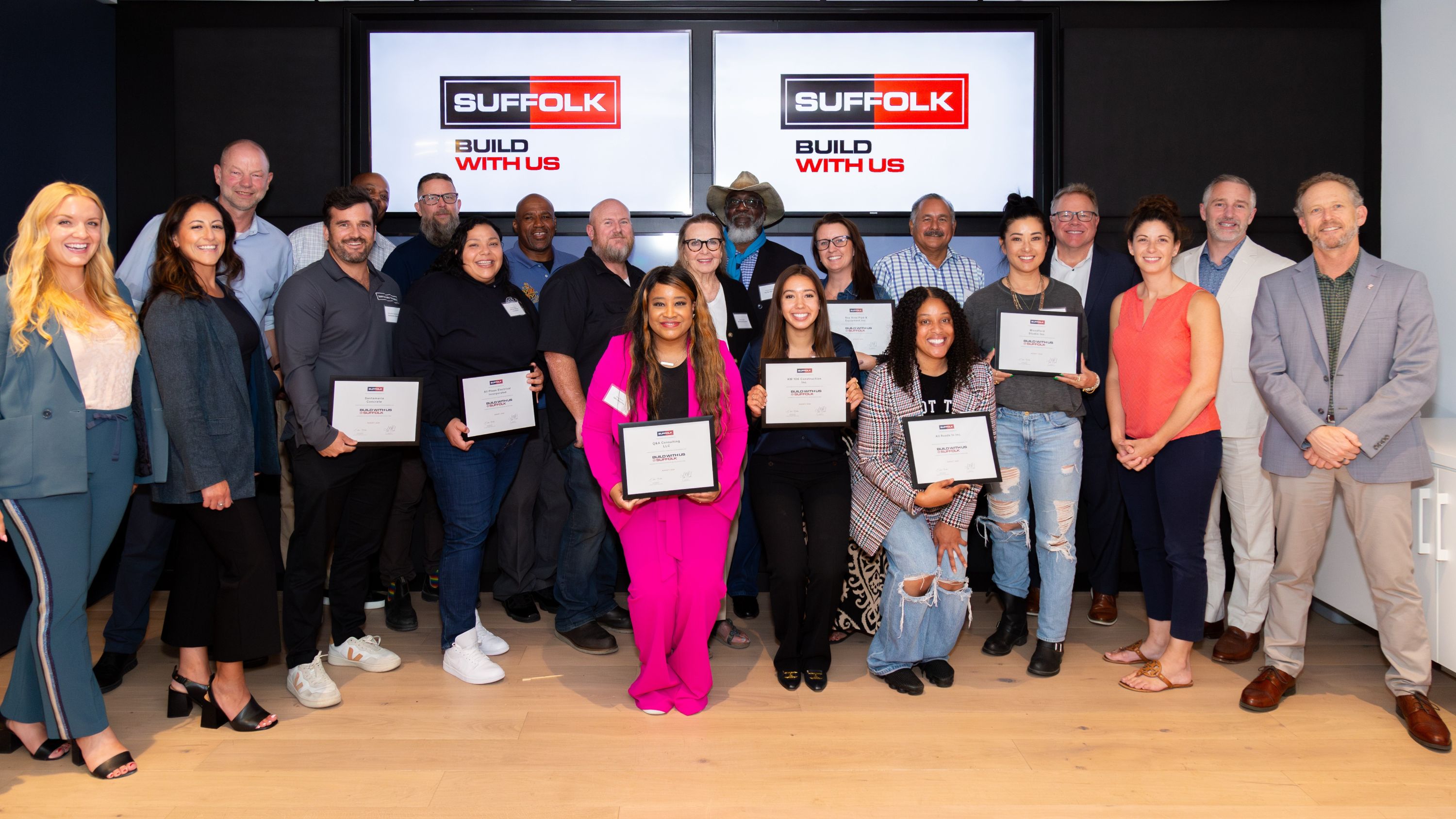 A diverse group of people stand together indoors, smiling and holding certificates. Behind them are two large screens displaying the Suffolk logo and the words Build With Us.
